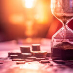 Sand running through an hourglass next to small stacks of coins in front of a setting sun