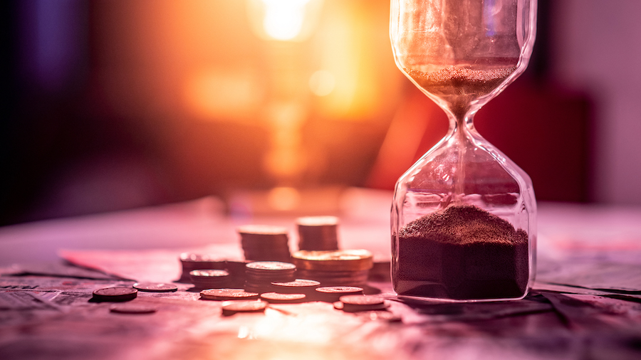 Sand running through an hourglass next to small stacks of coins in front of a setting sun