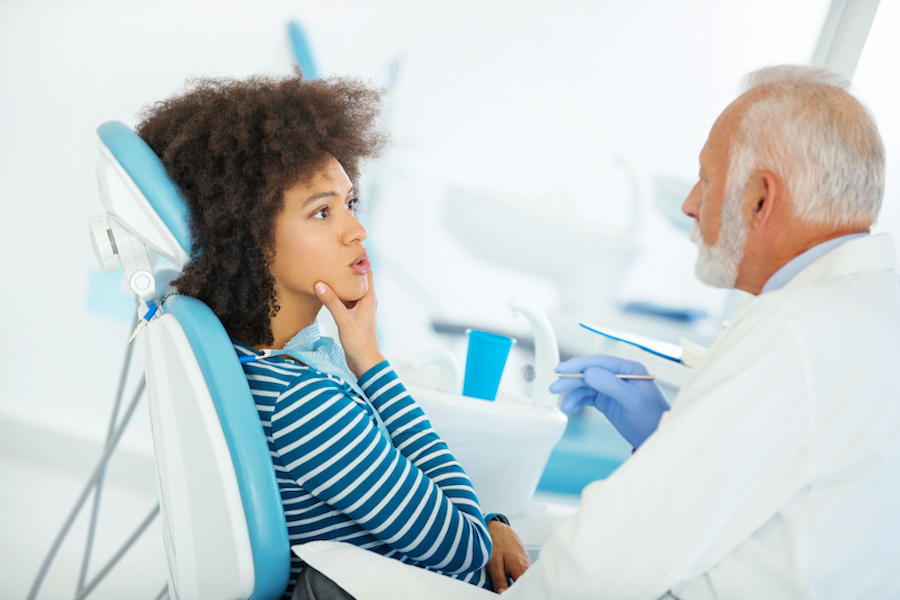 A Black woman touches her jaw as she sits in a dental chair having a consultation with her dentist