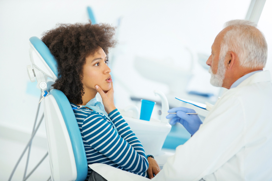 A Black woman touches her jaw as she sits in a dental chair having a consultation with her dentist, dentist in wynne