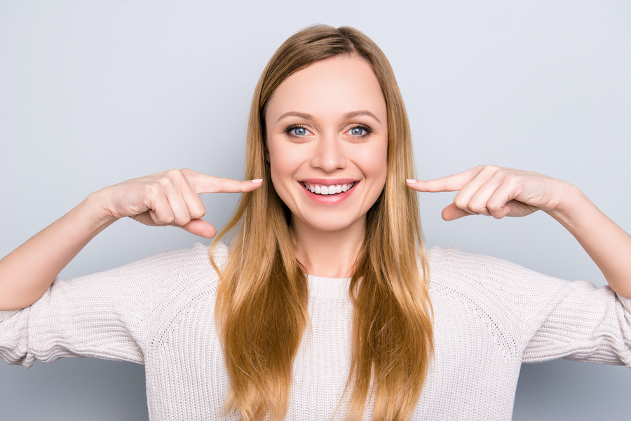 Blonde woman smiles and points at her teeth horizontally with her index fingers